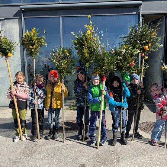 Eine Gruppe von Kindern, in warme Kleidung gehüllt, steht im Freien mit mit Grün und Früchten dekorierten Stöcken. Dahinter ist ein Gebäude mit Glasfenstern zu sehen.