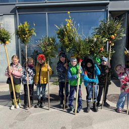 Eine Gruppe von Kindern, in warme Kleidung gehüllt, steht im Freien mit mit Grün und Früchten dekorierten Stöcken. Dahinter ist ein Gebäude mit Glasfenstern zu sehen.
