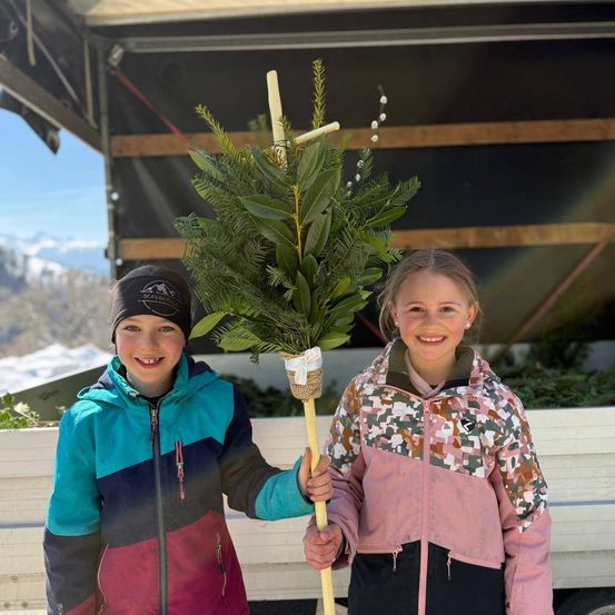 Zwei Kinder, lächelnd, stehen und halten eine dekorierte immergrüne Pflanze. Sie stehen in einer winterlichen Außenumgebung mit verschneiten Bergen im Hintergrund.