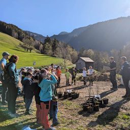 Eine Gruppe von Kindern und Erwachsenen versammelt sich in einer Bergregion. Sie umgeben kleine Topfpflanzen und lauschen einem uniformierten Mann.