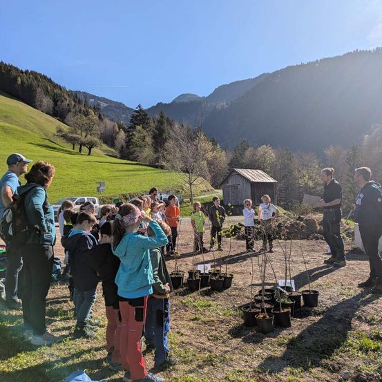 Eine Gruppe von Kindern und Erwachsenen versammelt sich in einer Bergregion. Sie umgeben kleine Topfpflanzen und lauschen einem uniformierten Mann.