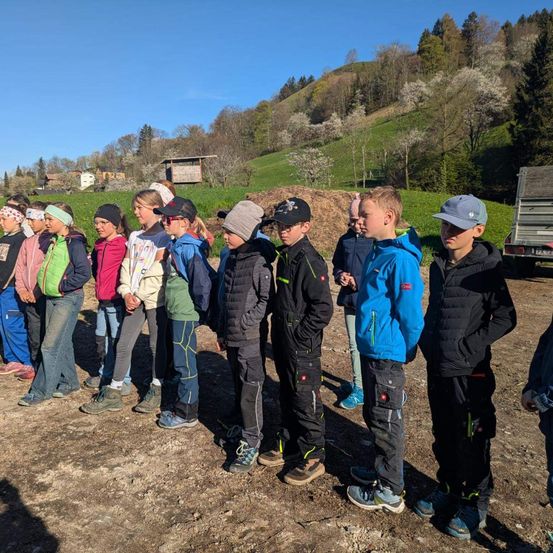 Eine Gruppe von Kindern steht in einer Reihe auf einem Feld aus Erde. Einige tragen Mützen, und einige tragen Schuhe. Das Feld hat einen grasbewachsenen Hang mit Bäumen. In der Ferne befindet sich ein Haus mit einem LKW dahinter.