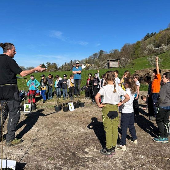 Eine Gruppe von Kindern in grünen und orangefarbenen Shirts steht um einen Mann herum, der sie in einem Feld mit Bäumen unterrichtet.