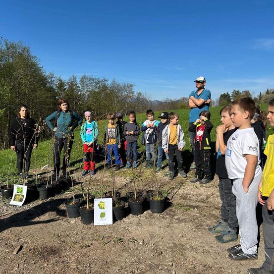 Eine Gruppe von Kindern und Erwachsenen steht um kleine Topfpflanzen in einem Feld unter blauem Himmel.