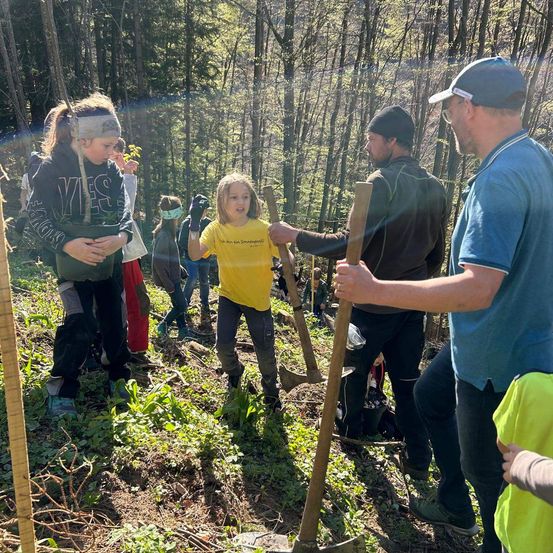 Eine Gruppe von Menschen pflanzt Bäume in einem Wald. Ein Mann gibt einer Frau in einem gelben Shirt Anweisungen. Dahinter hält eine weitere Person eine Schaufel.