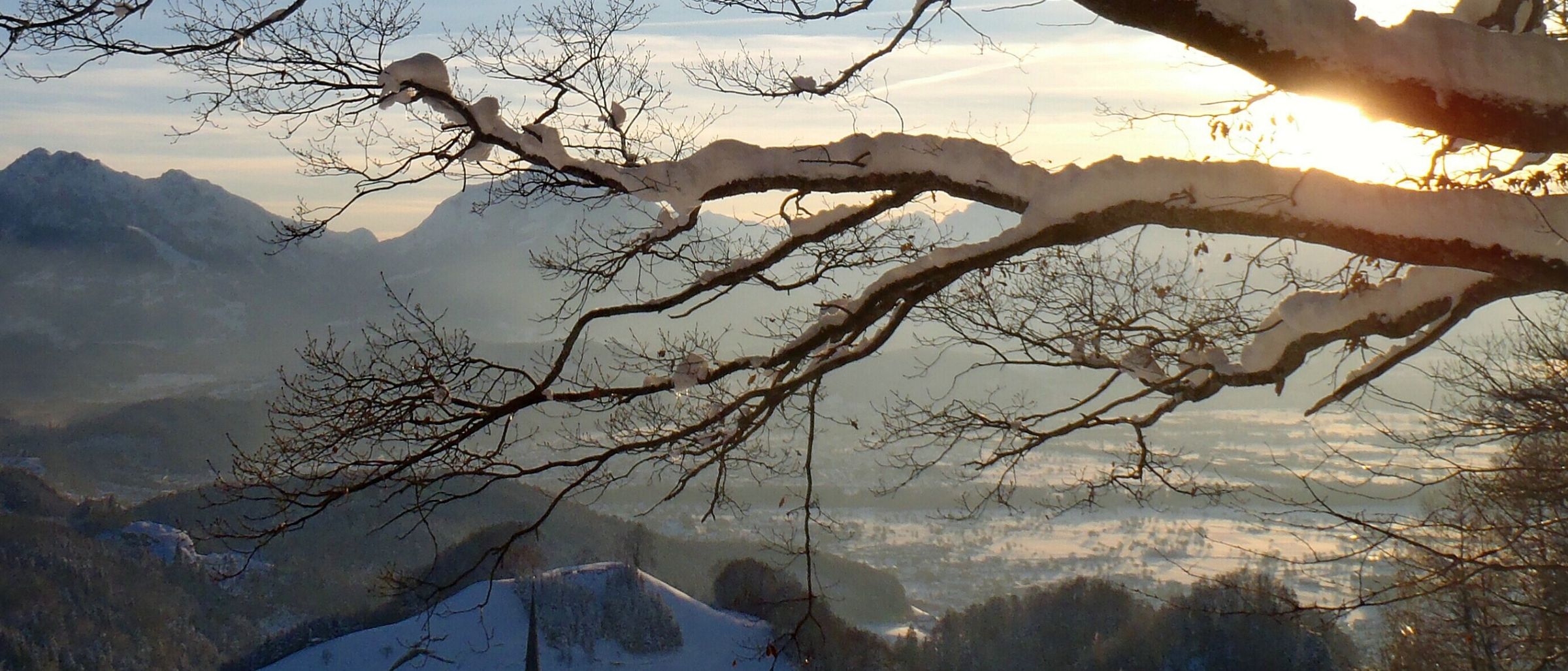 Schneebedeckte Berge, ein Dorf unten und ein Kirchturm, der zum Himmel reicht. Eine ruhige Winterlandschaft unter blauem Himmel.