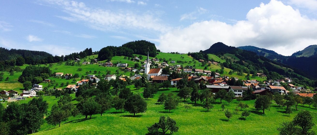 Ein Dorf am Hang mit einer Kirche, umgeben von sattgrünen Feldern, Bäumen und Bergen unter einem blauen Himmel mit Wolken.