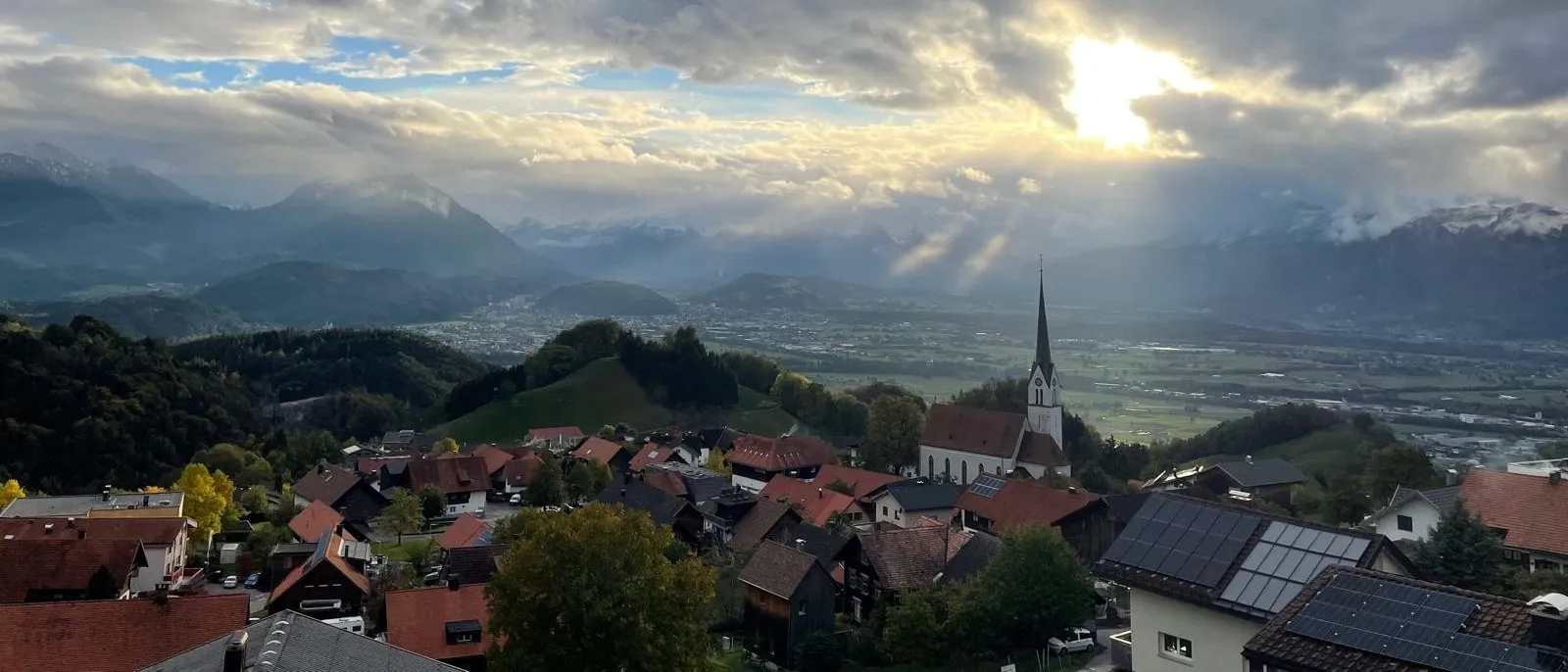 Ein Luftbild einer kleinen Stadt mit roten Dächern und einem Kirchturm, umgeben von grünen Hügeln und Bergen unter einem bewölkten Himmel.