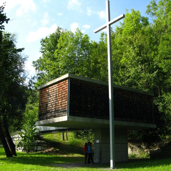 Eine moderne Kirche mit Betonaußerem und einem großen Kreuz davor, in einem Wald gelegen, mit zwei Personen, die in der Nähe stehen.