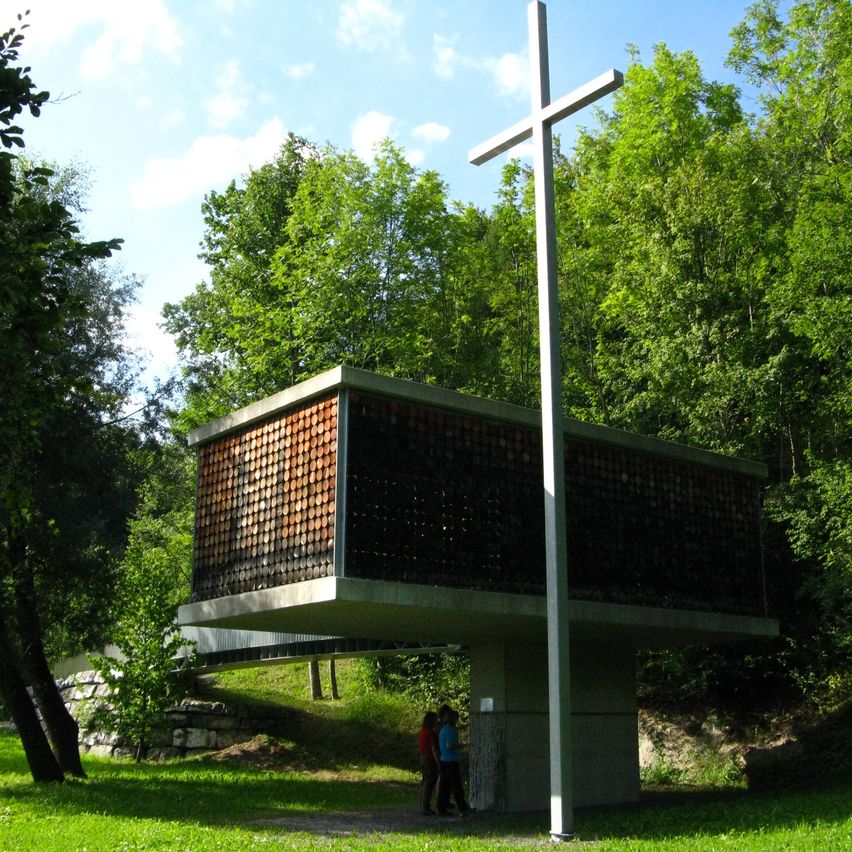 Eine moderne Kirche mit Betonaußerem und einem großen Kreuz davor, in einem Wald gelegen, mit zwei Personen, die in der Nähe stehen.