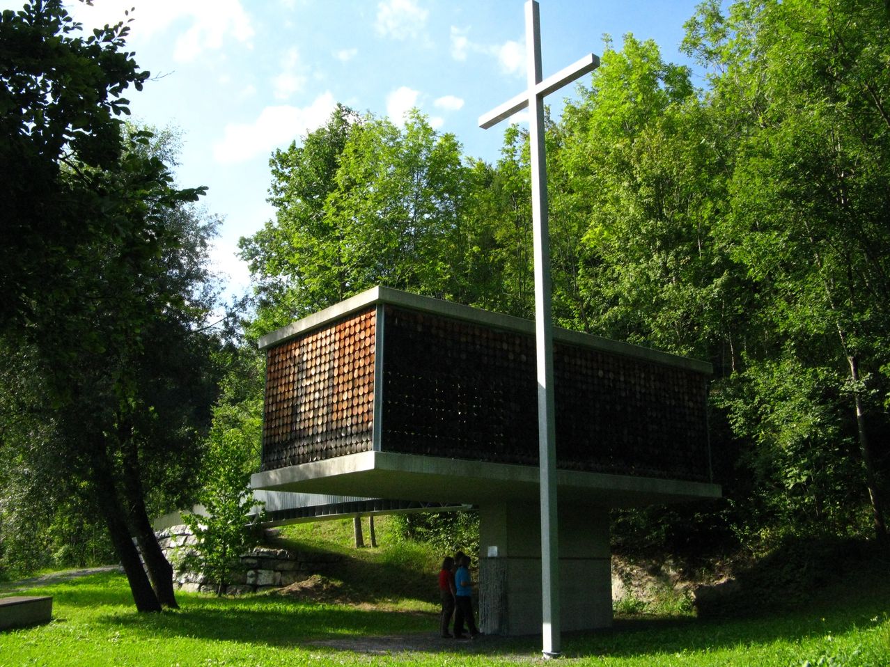 Eine moderne Kirche mit Betonaußerem und einem großen Kreuz davor, in einem Wald gelegen, mit zwei Personen, die in der Nähe stehen.