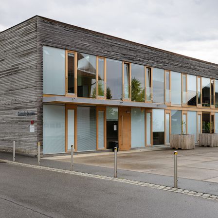 Ein modernes Gebäude mit einer Holzfassade und großen Glasfenstern. Auf der rechten Seite befinden sich zwei Holzpflanzer mit Pflanzen.