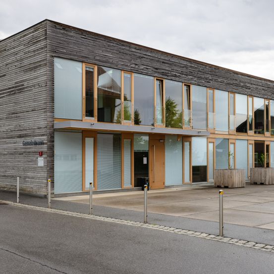 Ein modernes Gebäude mit einer Holzfassade und großen Glasfenstern. Auf der rechten Seite befinden sich zwei Holzpflanzer mit Pflanzen.