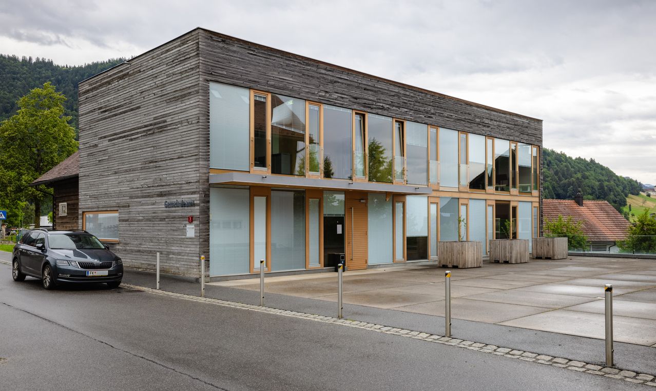 Ein modernes Gebäude mit einer Holzfassade und großen Glasfenstern. Auf der rechten Seite befinden sich zwei Holzpflanzer mit Pflanzen.