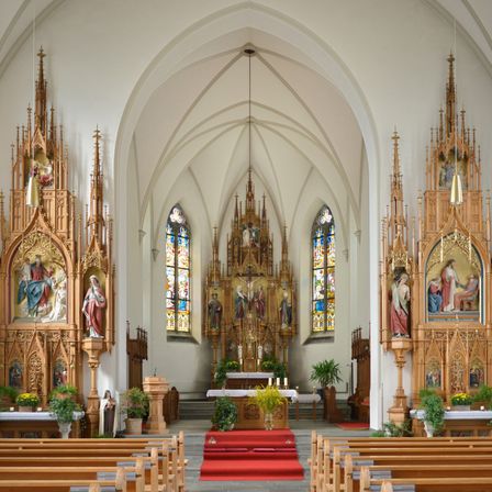Der Innenraum einer Kathedrale zeigt zwei kunstvoll verzierte Altäre, Buntglasfenster und einen roten Teppich, der zum Altar führt. Holzbänke sind auf beiden Seiten angeordnet.