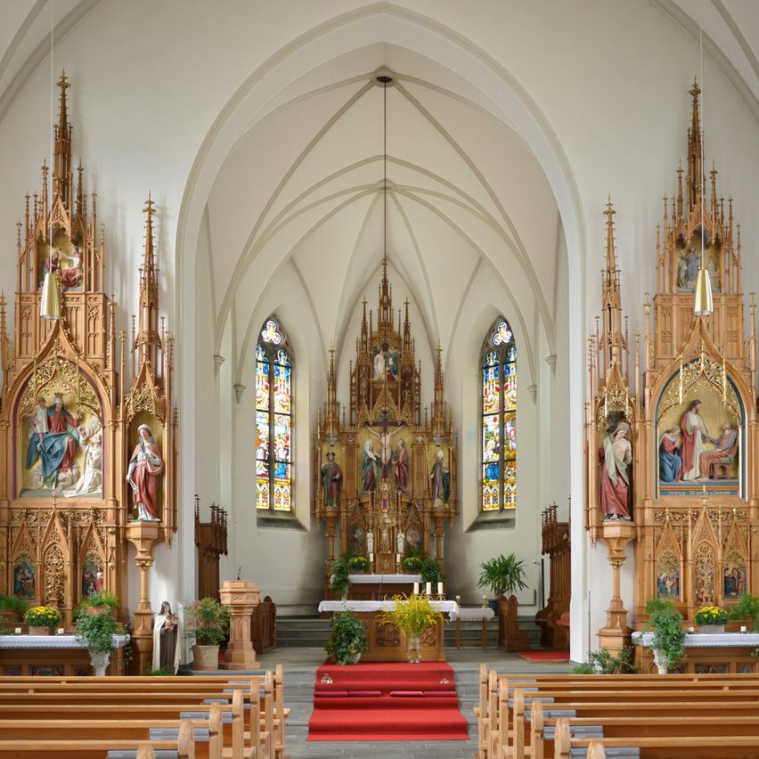 Der Innenraum einer Kathedrale zeigt zwei kunstvoll verzierte Altäre, Buntglasfenster und einen roten Teppich, der zum Altar führt. Holzbänke sind auf beiden Seiten angeordnet.