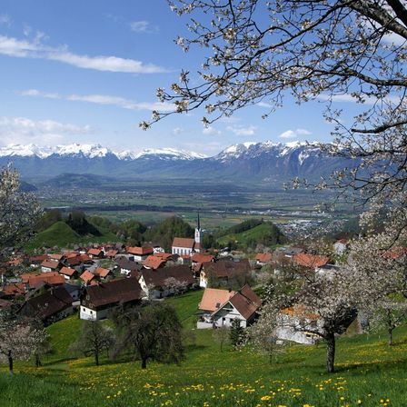 Ein malerisches Dorf mit roten Dächern, eingebettet in einem üppigen grünen Tal, mit blühenden Bäumen und Bergen im Hintergrund unter einem blauen Himmel.