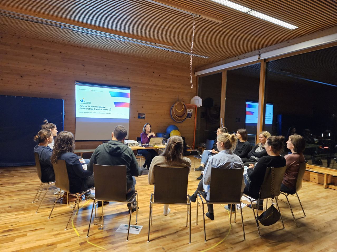 Eine Gruppe von Frauen sitzt in Stühlen um einen Tisch in einem Konferenzraum. Eine Frau spricht vor einer Projektionsleinwand.