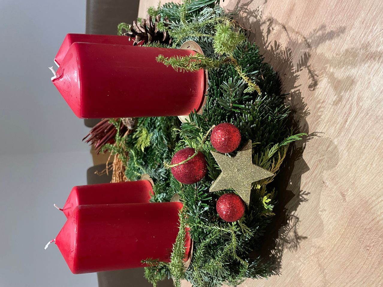 Ein Kranz mit roten Kerzen und Weihnachtskugeln, mit einem goldenen Stern und Tannenzapfen, steht auf einer Holzoberfläche.