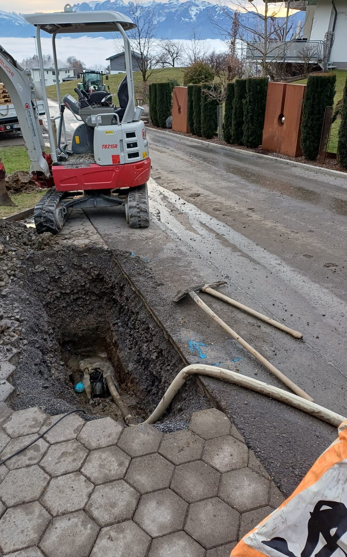 Ein roter und weißer TB215R-Bagger gräbt ein Loch in der Straße. Zwei Hämmer liegen auf dem Boden. Das Loch ist mit Erde gefüllt und hat einen blauen Gegenstand darin.