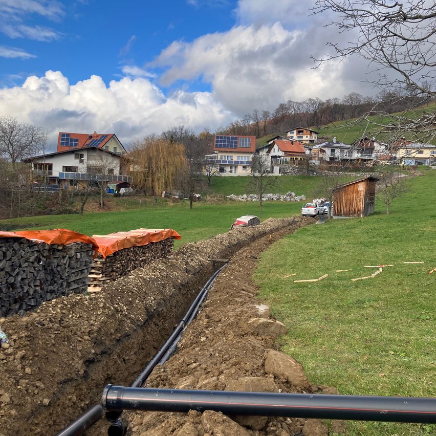 Ein offener Graben für die Verlegung von Rohrleitungen liegt in einem Grasfeld mit Häusern mit Solarmodulen im Hintergrund.
