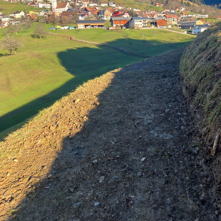 Luftaufnahme eines ländlichen Dorfes mit Häusern, Bäumen und einem grasbewachsenen Feld. Der Schatten eines Hügels fällt über das Feld.