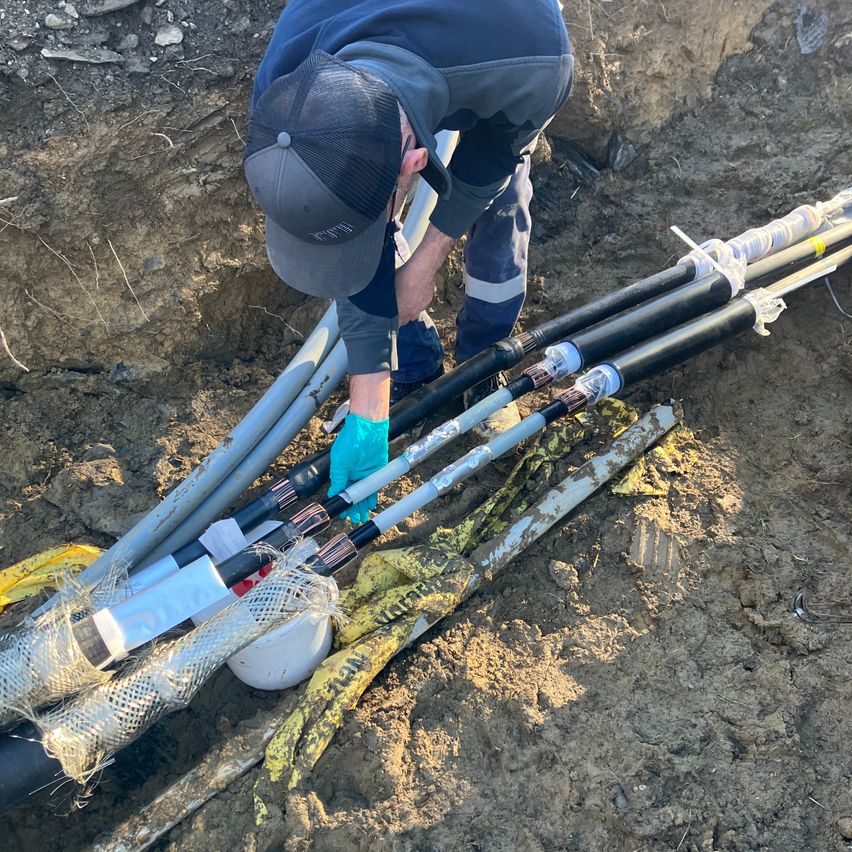 Ein Arbeiter in einer Kappe und Handschuhen repariert Kabel im Boden. Die Kabel sind umwickelt und mit Klebeband gesichert. Das Gebiet ist mit Erde bedeckt.