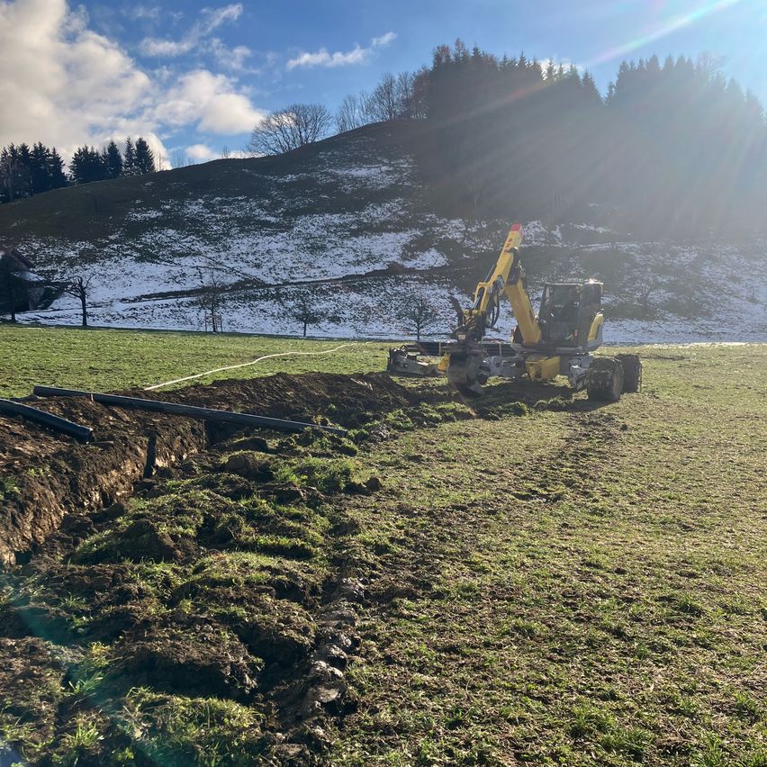 Ein Baufahrzeug gräbt einen Graben in einem Grasfeld unter klarem Himmel. Im Hintergrund befinden sich schneebedeckte Berge.