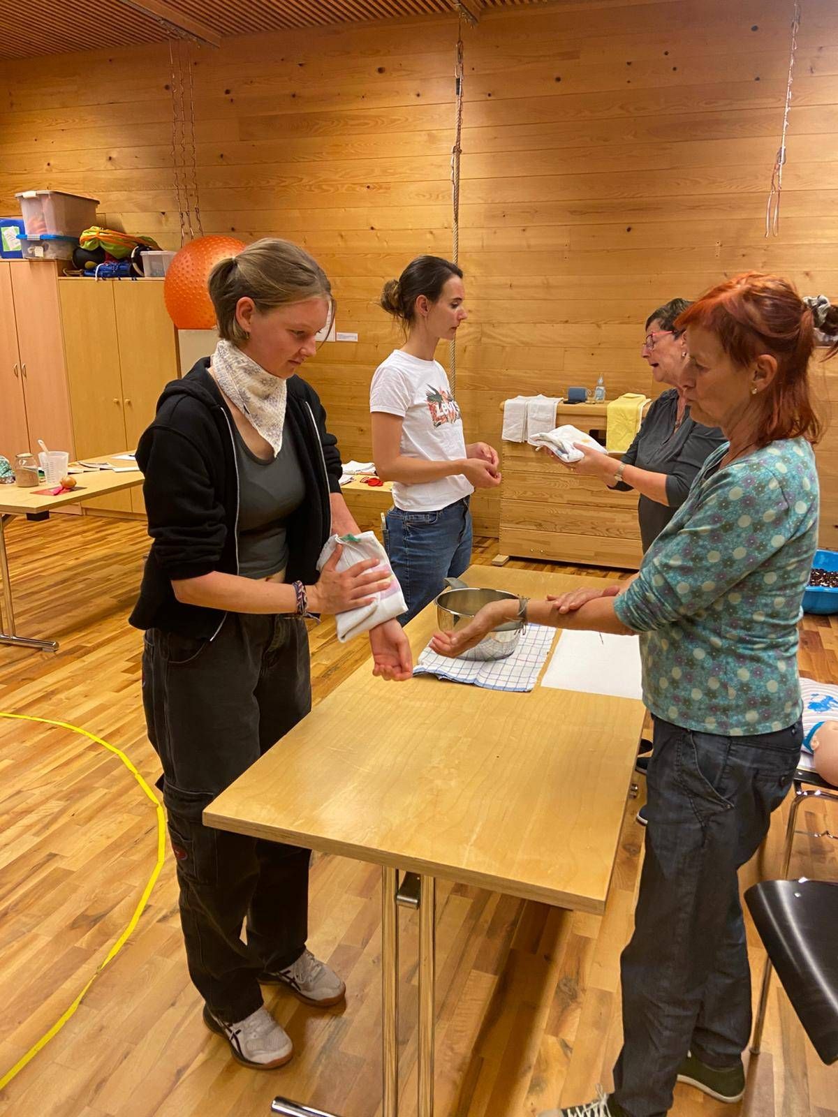 Eine Gruppe von Frauen ist um einen Holztisch versammelt, wobei eine Frau eine Schüssel hält und eine andere etwas erhält. Sie befinden sich in einem Raum mit Holzwänden und einem Holzboden.