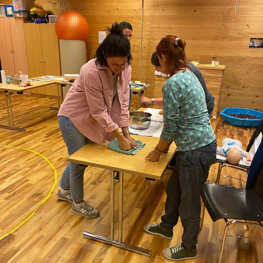 Drei Frauen befinden sich in einem Raum mit Holzwänden und bereiten einen Tisch vor. Eine Frau steht und wischt ein Tuch, während eine andere neben ihr steht. Ein Baby liegt in einem Stuhl in der Nähe.