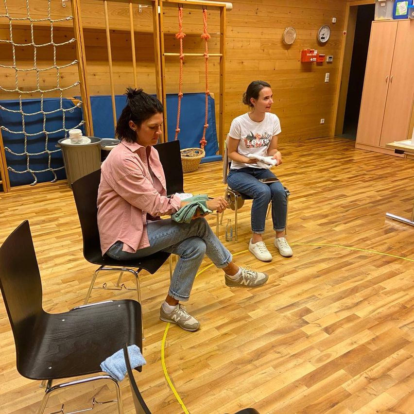 Zwei Frauen sitzen in Stühlen in einem Raum, eine trägt ein rosa Shirt und die andere ein weißes. Sie scheinen miteinander zu sprechen. Der Raum hat Holzböden und -wände.