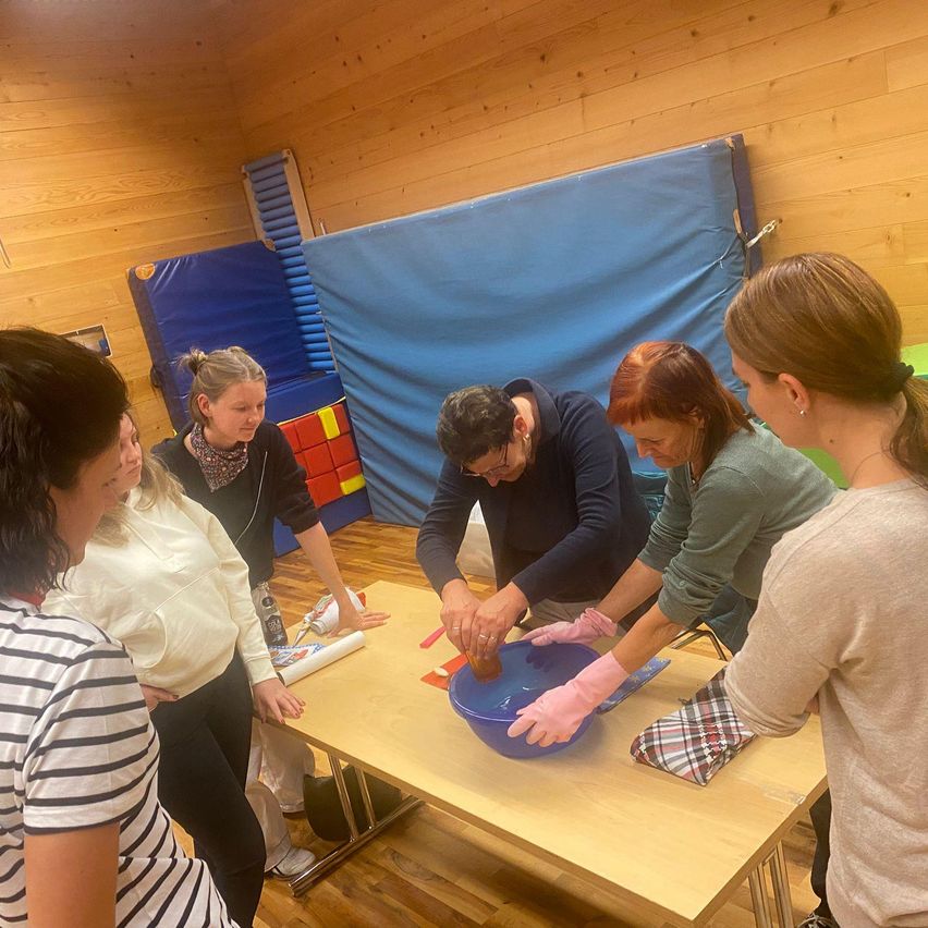 Mehrere Frauen arbeiten in einem Raum mit Holzwänden an einem Projekt. Sie stehen um einen Tisch mit einer blauen Schüssel herum, tragen Handschuhe und bereiten etwas vor.