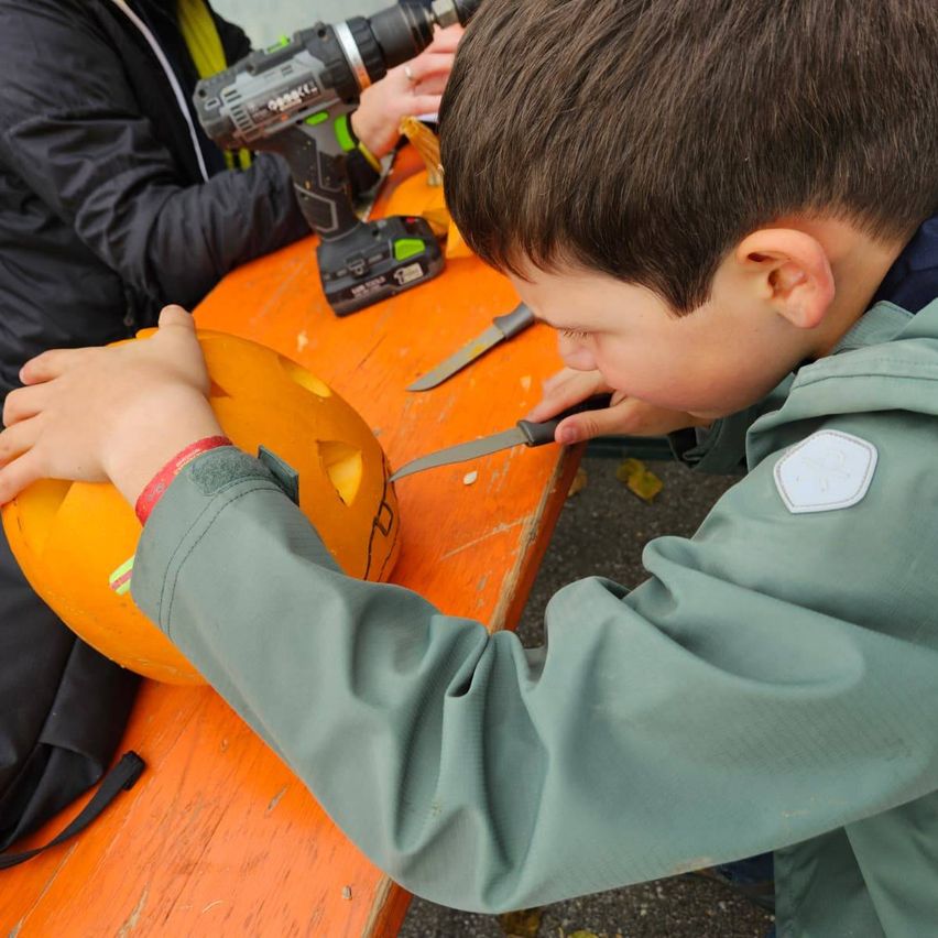 Ein Junge schnitzelt eine Kürbis mit einem Messer, während eine Frau in der Nähe eine Bohrmaschine benutzt.