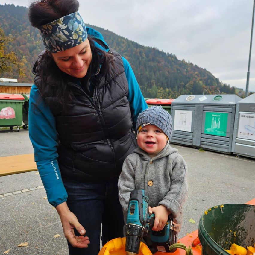 Eine Frau und ein junger Junge halten eine Bohrmaschine und stehen neben einer Kürbis. Sie lächeln und scheinen an einer Aktivität beteiligt zu sein. Hinter ihnen befinden sich Mülleimer und eine Berglandschaft.