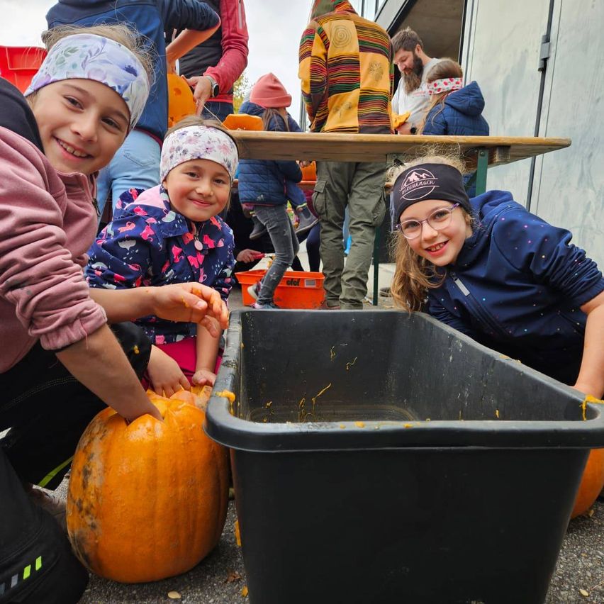 Drei junge Mädchen lächeln, während sie draußen Kürbisse schnitzen. Ein schwarzer Mülleimer ist in der Nähe, und einige Erwachsene sind im Hintergrund.