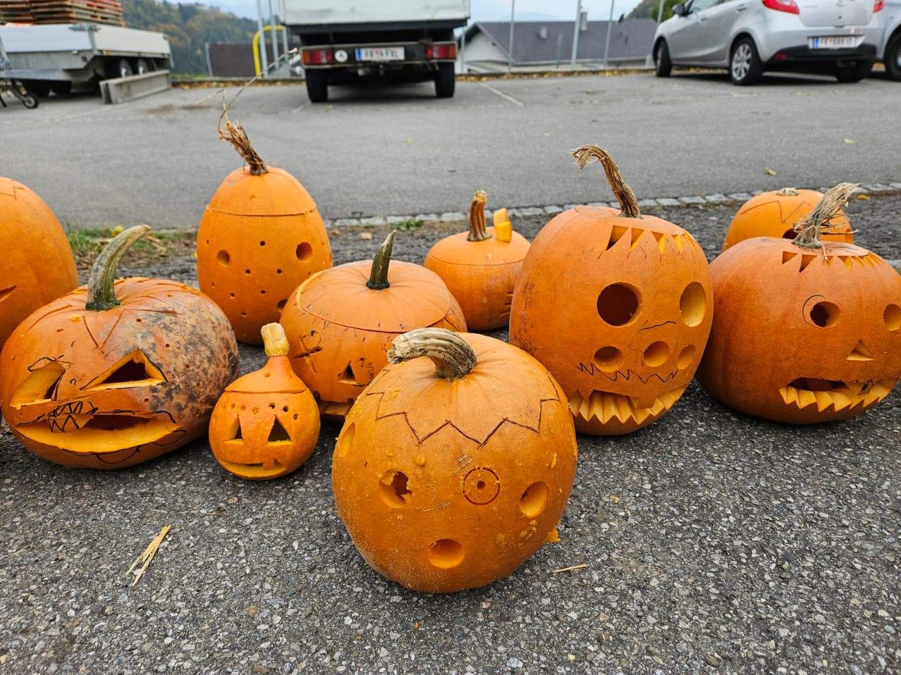 Verschiedene geschnitzte Kürbisse, einige mit Augen und Zähnen, sind im Freien auf einem Parkplatz ausgestellt. Im Hintergrund sind zwei Fahrzeuge geparkt, ein LKW und ein Auto.
