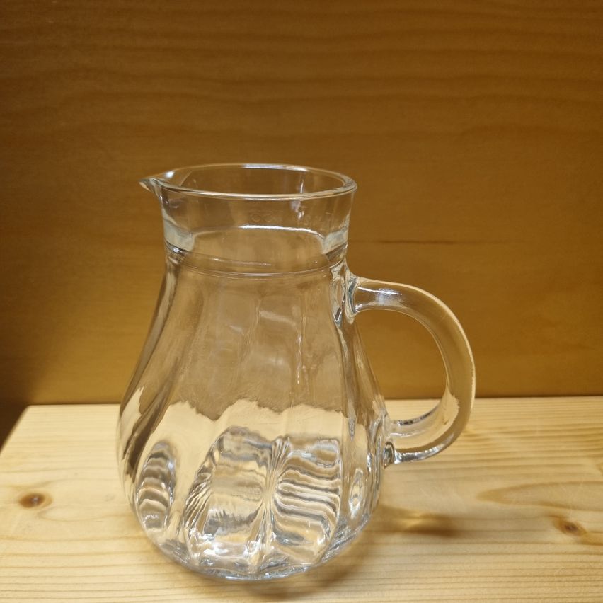 Ein leerer klarer Glaskrug mit einem gebogenen Griff steht auf einem Holztisch. Der Krug hat ein dekoratives Muster am Boden.