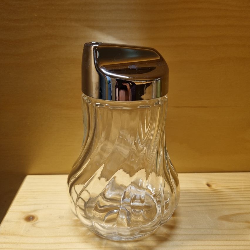 Eine klare Glasflasche mit silbernem Deckel steht auf einem Holztisch. Die Flasche hat ein einzigartiges Design mit einer wellenförmigen Form. Die Reflexion der Flasche ist auf dem Deckel sichtbar.