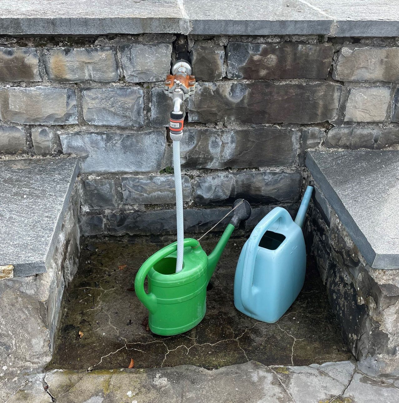 Zwei Gießkannen, eine grün und eine blau, stehen neben einem Außenhahn mit angeschlossenem Schlauch, in einem Betonbrunnen platziert.