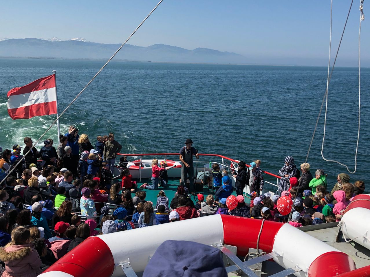 Ein Boot mit vielen Menschen auf dem Deck, einige stehen in der Nähe der Reling, eine Person hält ein Kind. Eine Flagge weht links. Im Hintergrund ein ruhiges Meer und Berge.