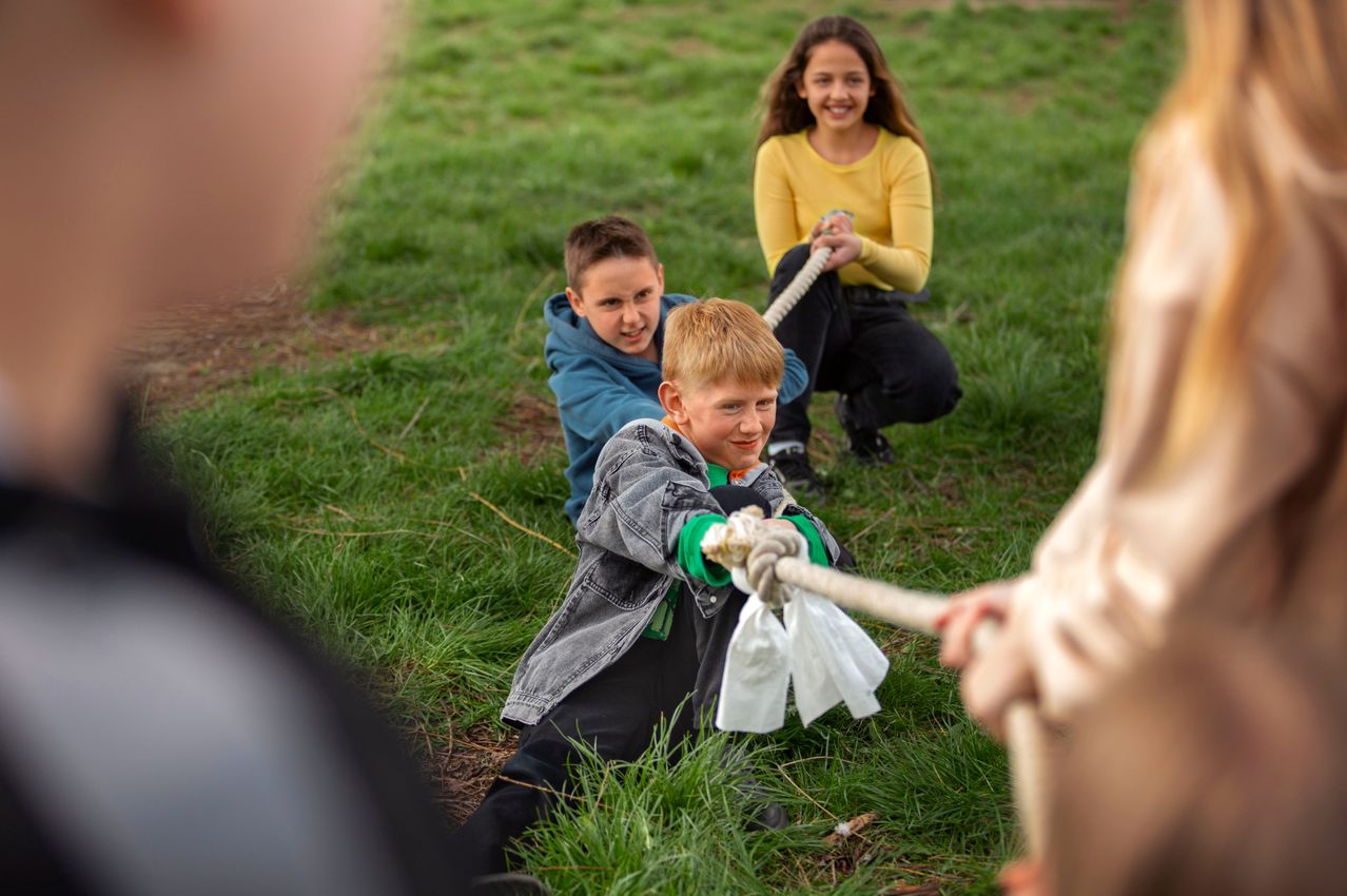 Drei Kinder spielen auf einer Grasfläche Tauziehen, eines zieht, eines zieht zurück und ein Mädchen beobachtet.