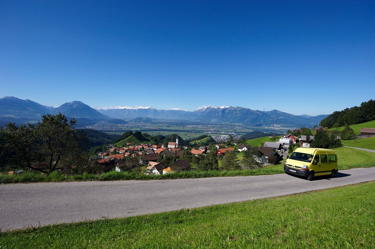 Ein gelber Van fährt eine Straße hinunter, mit Blick auf ein Dorf, Berge und einen blauen Himmel.