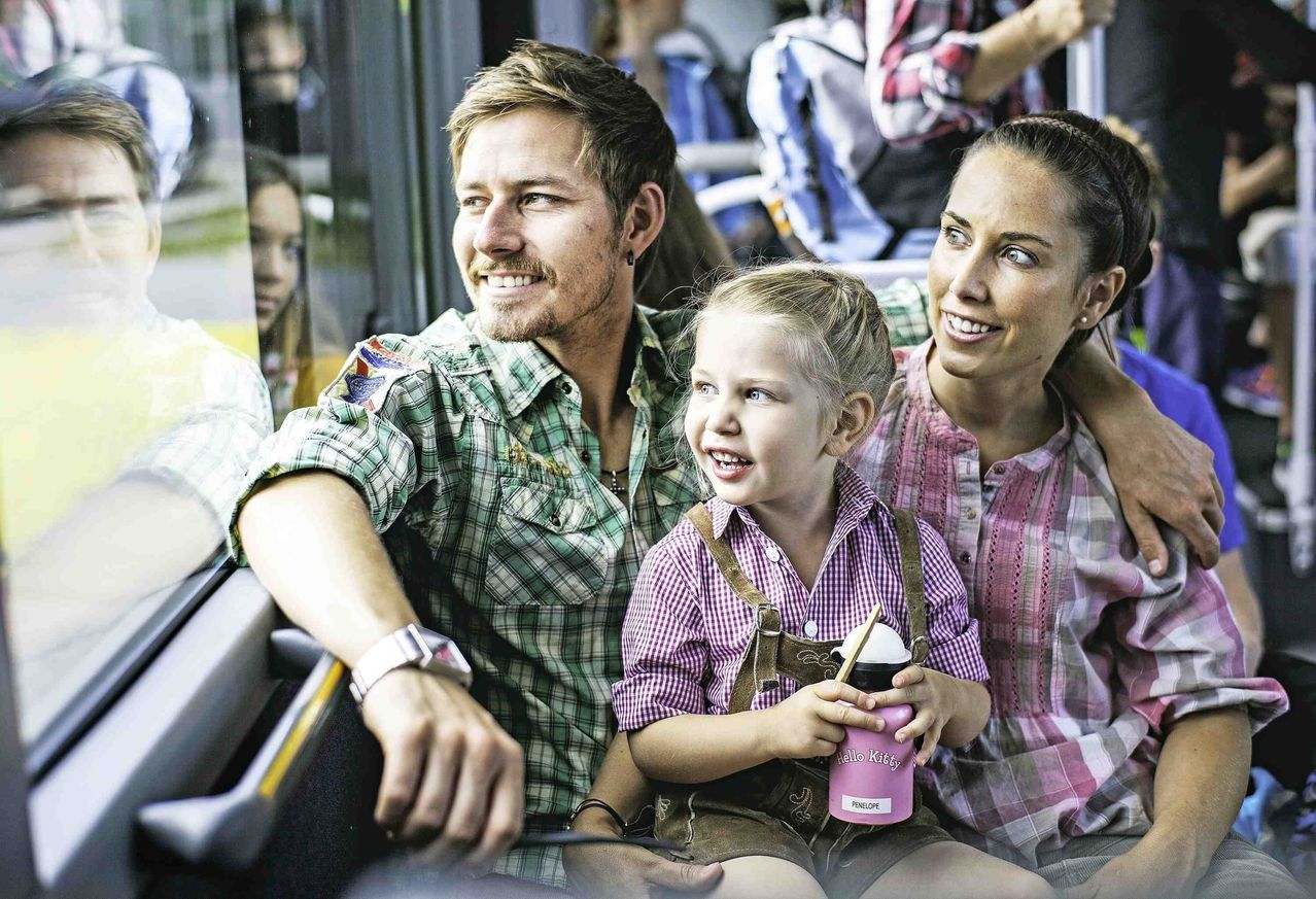 Eine dreiköpfige Familie sitzt in einem Bus. Der Mann lächelt und trägt ein kariertes Hemd, die Frau neben ihm trägt ein rosa Hemd und das Kind hält einen lila Becher.