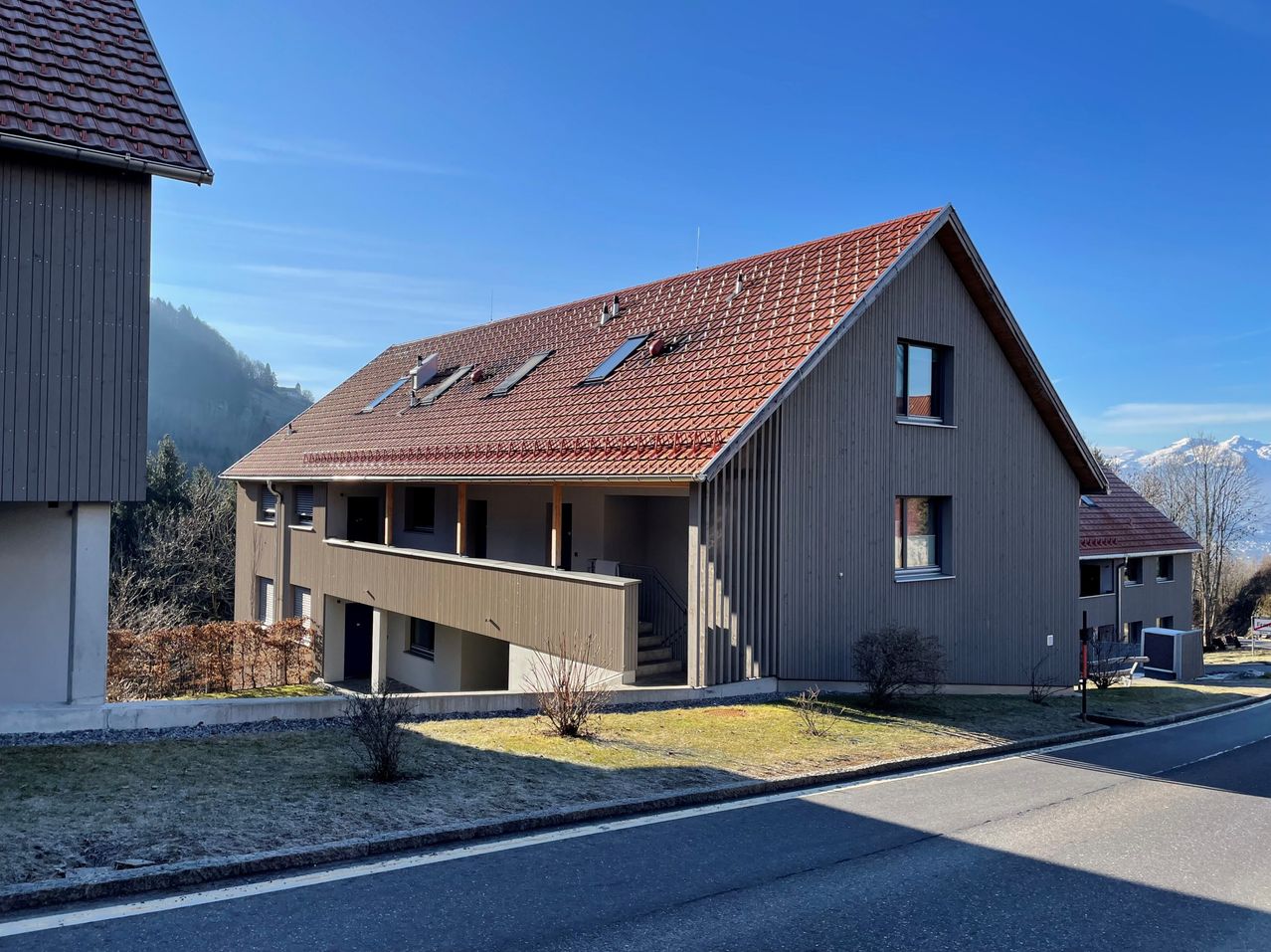 Ein modernes Haus mit rotem Ziegeldach und Solarpanels, umgeben von Rasen und Büschen, mit einer Straße in der Nähe.