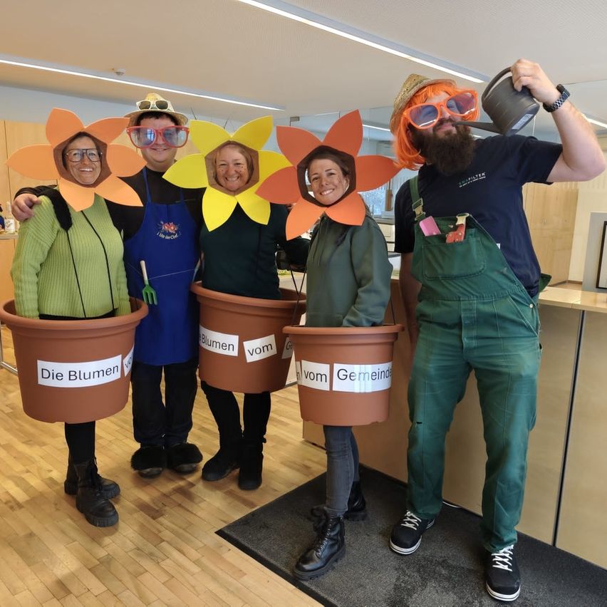 Fünf Menschen in Gartenkostümen posieren drinnen. Jeder hält einen Blumentopf mit einem Etikett. Ein Mann mit roter Perücke hält eine Gießkanne. Dahinter steht eine Holztheke.