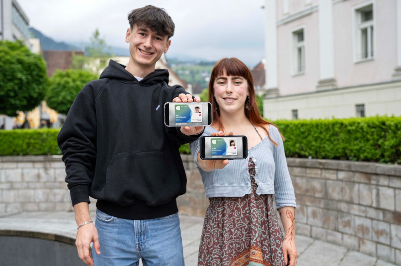Ein junger Mann und eine junge Frau stehen draußen, lächeln und zeigen ihre digitalen Ausweise auf ihren Handys. Sie sind lässig gekleidet, und der Mann trägt eine schwarze Kapuzenjacke. Hinter ihnen steht ein Gebäude mit Fenstern und Grünpflanzen.