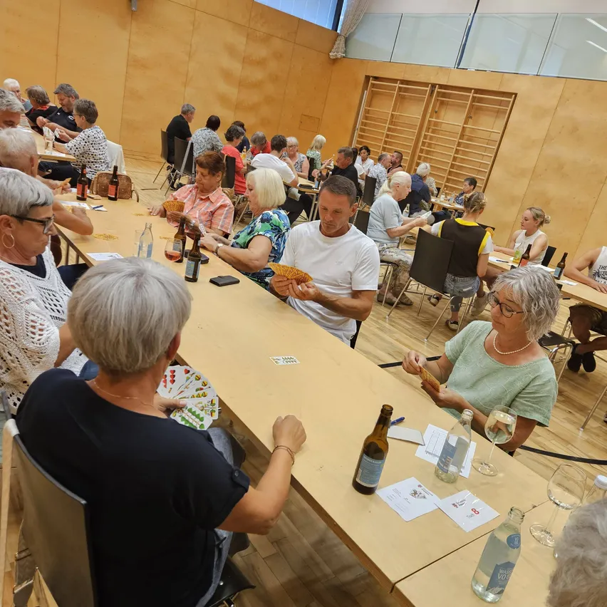 Eine Gruppe älterer Menschen sitzt um einen langen Tisch herum und spielt Karten. Der Raum hat Holzböden und Wände, und es gibt Flaschen und Gläser auf dem Tisch.