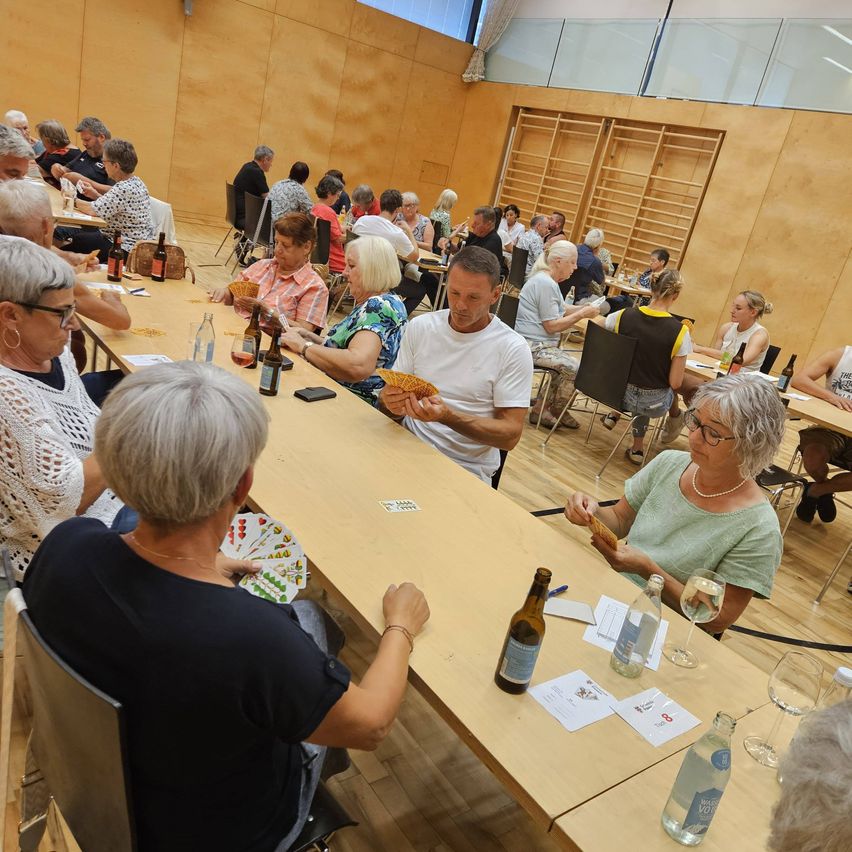 Eine Gruppe älterer Menschen sitzt um einen langen Tisch herum und spielt Karten. Der Raum hat Holzböden und Wände, und es gibt Flaschen und Gläser auf dem Tisch.