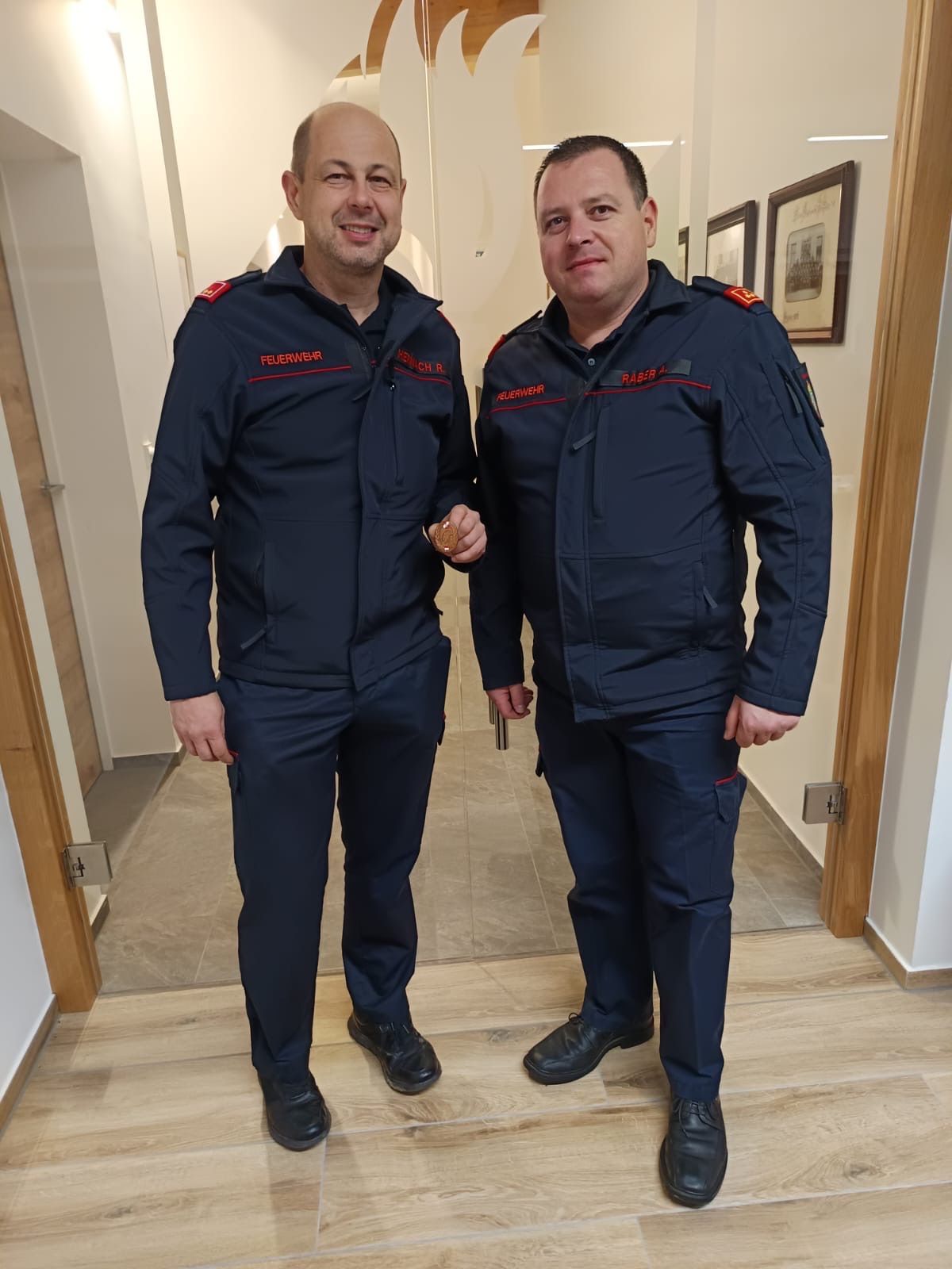 Two men in blue uniforms with red stripes and the word 'Feuerwehr' stand in a hallway, smiling. The man on the left holds a coin, and both wear black shoes. Behind them are wooden walls with picture frames.