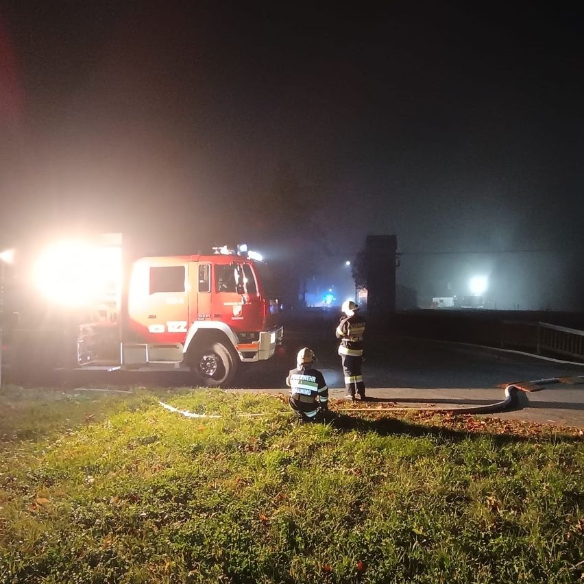 Two firefighters are standing by a red fire truck with its lights on. They are near a grassy area at night.